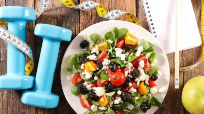 A plate of colorful salad with greens, tomatoes, and cheese, surrounded by dumbbells and a measuring tape.