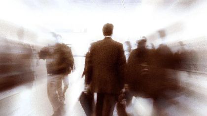 A blurred image of a man in a suit walking through a busy airport with rushing travelers around him.