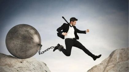 A man in a suit leaps away from a large metal ball and chain over rocky terrain, symbolizing freedom or struggle.