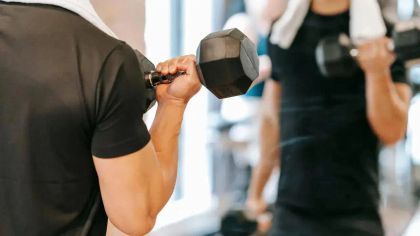 A person lifting a dumbbell in a gym, with a towel around their neck and a mirror reflecting their workout.