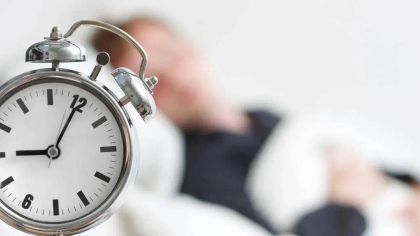 A close-up of an alarm clock showing 7:00, with a blurry figure sleeping in bed in the background.