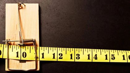 A mousetrap on a black surface, with a yellow measuring tape extending across it, showing measurements up to 14 inches.