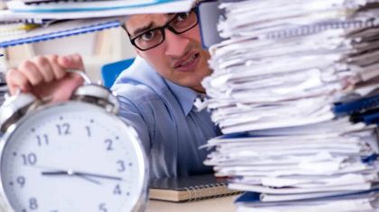 A stressed man in glasses reaches for a clock, surrounded by tall stacks of paperwork on his desk.