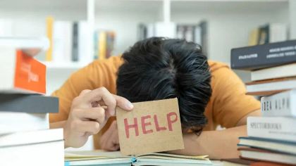 A frustrated person with their head on a book holds up a card reading "HELP" in a study environment.