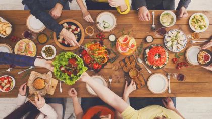 Aerial view of a diverse feast on a wooden table, with hands reaching for salads, sushi, and various dishes.