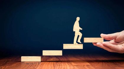 A hand holds a wooden block as a silhouette of a person climbs a staircase made of blocks against a dark background.