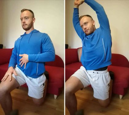 A man in a blue hoodie and shorts demonstrates a stretching exercise while seated on a floor with a red couch in the background.