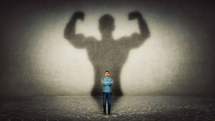 A person stands confidently with their arms crossed, casting a large shadow of a muscular figure on the wall behind.