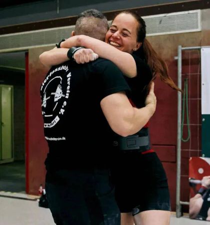 Two athletes embrace joyfully after a successful lift, both wearing black t-shirts in a training gym setting.