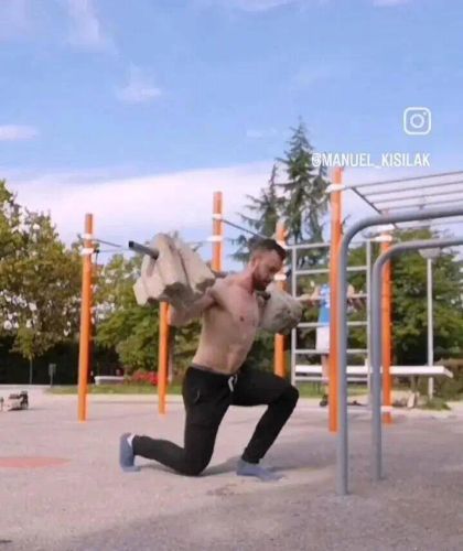 A shirtless man performs a lunge while carrying a sandbag in an outdoor park gym with workout equipment in the background.