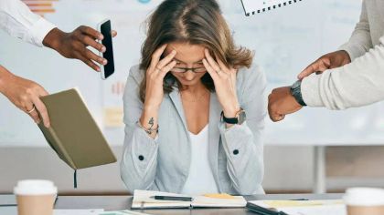 A woman at her desk looks stressed, surrounded by hands holding phones and notebooks, indicating a busy work environment.