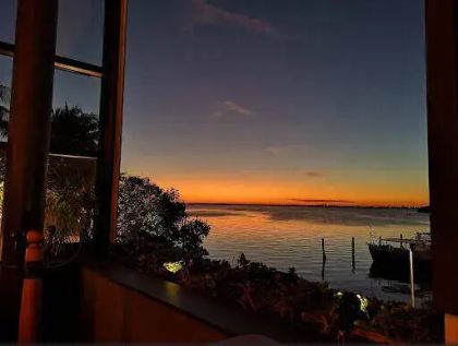 View of a serene sunset over water, framed by a window, with silhouettes of trees and a distant dock.