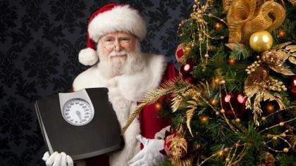 Santa Claus, dressed in traditional attire, stands beside a decorated Christmas tree holding a scale.