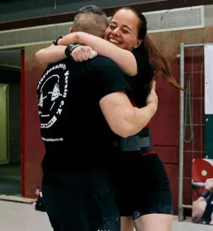 Two individuals embracing joyfully in a gym setting, both wearing black shirts and looking happy.