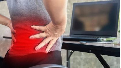 Person holding lower back in pain, seated near a laptop on a desk, indicating discomfort or strain.