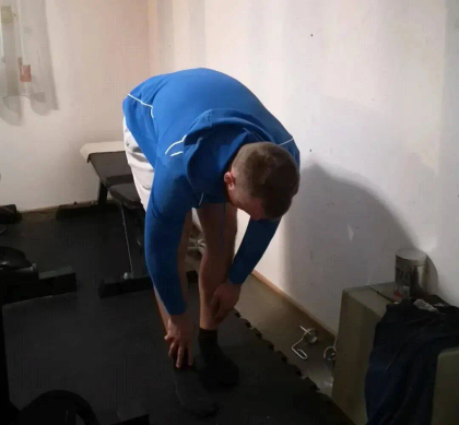 A person in a blue jacket bends down to stretch their leg in a home gym setting.